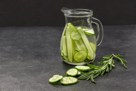 Cucumber infused water in glass jug. Sprig of rosemary on table. copyspace. Black background.の写真素材
