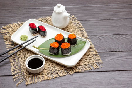 Nori sushi with caviar, black chopsticks on white plate. Soy sauce in bowl. White grave boat. Burlap napkin. copyspace. Dark wooden background. top view.の写真素材