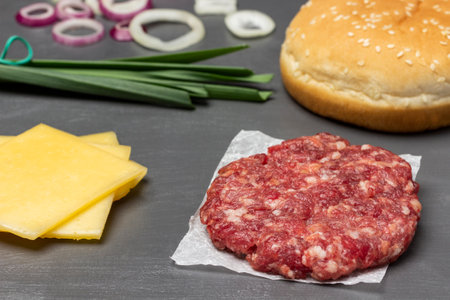 Sliced cheese, raw burger on paper, sesame bun and green onion on table. dark background. close up.の写真素材