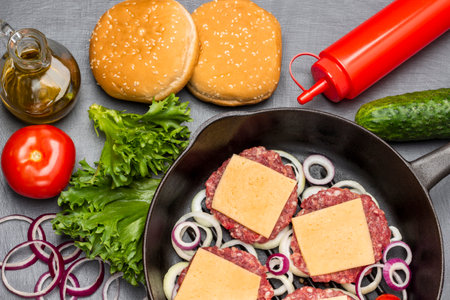 Buns with sesame green salad, tomato, cucumber and oil in glass bottle. Raw beef burgers with cheese and chopped onions in frying pan. flat lay. dark background.の写真素材
