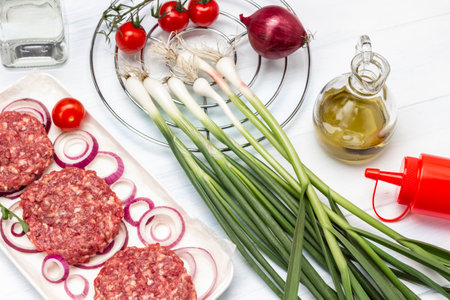 Green onions and garlic, tomatoes on branch and oil in glass bottle. Raw beef burgers with chopped onions on white plate. top view. white backgroundの写真素材