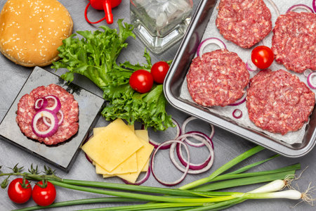 Green onions, cheese, water bottle. Buns with sesame seeds, green salad and tomatoes on table. Raw beef burgers in frying pan. flat lay. dark background.の写真素材