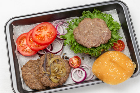 Fried burger bun on lettuce leaf, fried burger with cheese and fried onions, chopped tomatoes and onions on baking tray. flat lay. white backgroundの写真素材