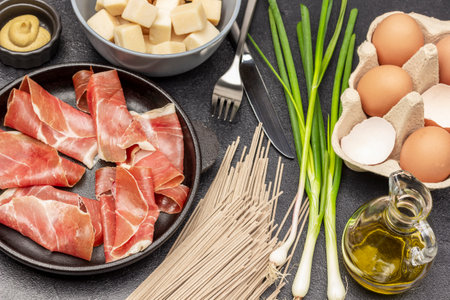 Dried ham in frying pan. Noodles, green onions on the table. Cheese and mustard in bowls. Eggs in box. top view. black backgroundの写真素材