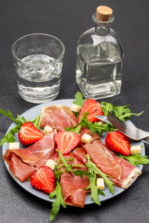 Spanish jamon with cheese, strawberries and arugula on gray plate. Fork and knife on plate. Glass bottle and glass of water. top view. black backgroundの写真素材