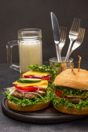 Cheeseburger with greens on bun with sesame seeds. Glass mug with beer. Knife and fork in tin. top view. black backgroundの写真素材
