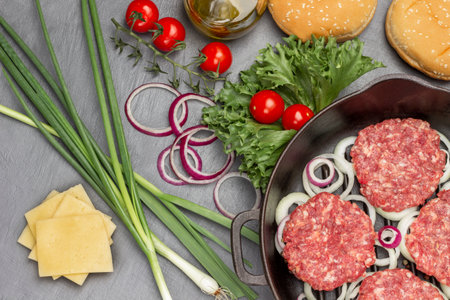 Raw burgers with onions in a frying pan. Cheese, green onions, tomato sprigs and lettuce on the table. flat lay. gray backgroundの写真素材