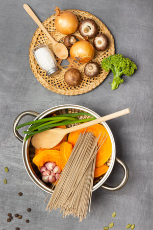 Noodles, pumpkin, chickpeas garlic and green stems in saucepan. Onions, mushrooms, salt shaker and cabbage on wicker plate. flat lay. gray backgroundの写真素材