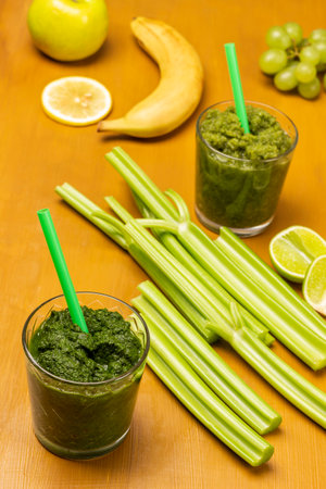Green smoothie and straw in glasses. Celery stalks and banana on table. Bright mustard background. top viewの写真素材