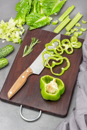 Half a green pepper, chopped green pepper, kitchen knife on cutting board. Two cucumbers, lettuce leaves and celery stalks on the table. flat lay. gray background.の写真素材