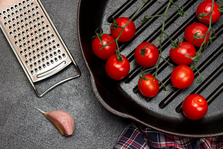 A sprig of cherry tomatoes in a frying pan. Grater and cloves of garlic on table. flat lay. Black background.の写真素材