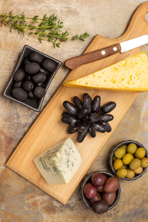 Two pieces of cheese and knife on the board. Green, red and black olives in bowls. Sprig of thyme and a bunch of black grapes. flat lay. Rusty metal background.の写真素材