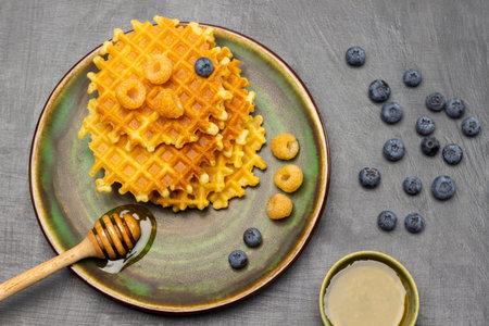 Wooden honey dipper, berries and waffles on green plate. Green bowl with honey and blueberries on table. flat lay. gray background.の写真素材