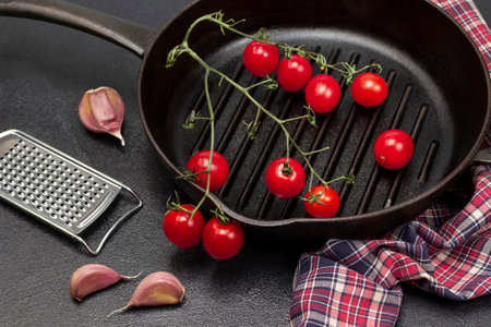 Garlic cloves and grater on the table. Sprig of red tomatoes in pan on napkin. top view. Black background.の写真素材