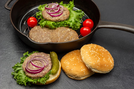 Grilled burger, pickle, onion and lettuce on round bun. Buns on the table. Burger, tomatoes and lettuce in frying pan. Black background.の写真素材