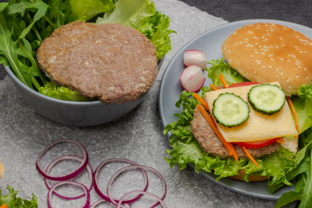 Beef burger with greens in bowl. Cheeseburger with cucumber, greens and carrots. Chopped onions on table. Black background.の写真素材