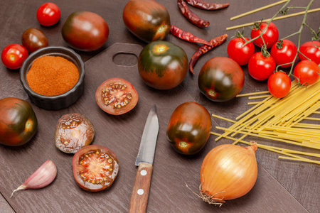 Kitchen knife and chopped tomatoes on cutting board. Dry paprika in bowl, pasta and whole tomatoes on table. top view. brown background.の写真素材