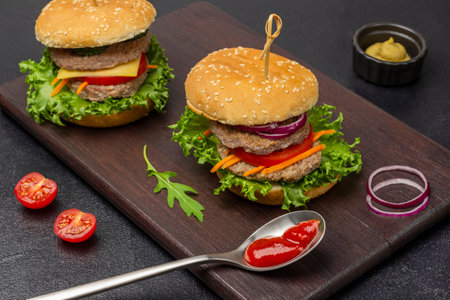 Two beef burgers with greens, chopped onions and carrots on board. Tomato sauce in spoon. Black background.の写真素材