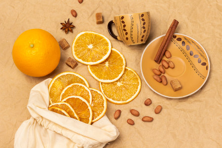 Coffee cup with saucer. Cinnamon stick and nuts. Slices of dried orange in fabric bag. fresh orange. flat lay. Beige paper background.の写真素材