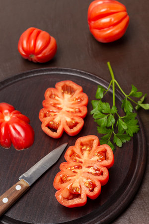 Halves of red tomatoes and a knife on a cutting board. Whole tomatoes on the table. top view. brown background.の写真素材