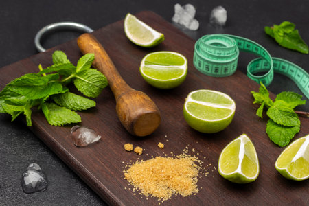 Lime halves, mint sprigs, wooden pestle and measuring tape on a board. Pieces of ice on the table. top view. Dark wooden background.の写真素材