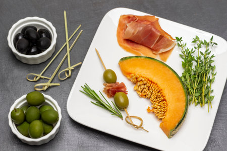 Two green olives and jamon on skewer. Slice of melon and sprigs of thyme on plate. Black and green olives in bowls. flat lay. gray background.の写真素材