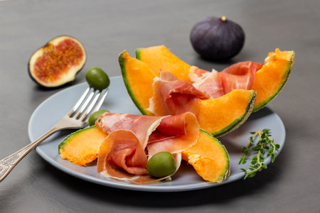 Green olives on a fork, jamon on a slice of melon, sprigs of thyme on a gray plate. Fresh figs on the table.の写真素材