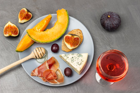 Honey stick and cheese, brown olives, jamon and melon on gray plate. Glass of wine and halves of figs on table. flat lay. gray background.の写真素材