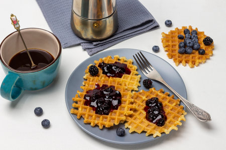 Waffles with jam and fork on gray plate. A blue cup of coffee and a coffee pot on gray napkin. top view. white background.の写真素材