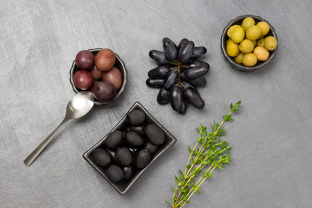 Green, red and black olives in bowls. Sprig of thyme and a bunch of black grapes. copyspace. flat lay. gray background.の写真素材