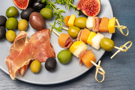Skewers with olives, cheese, and melon on a gray plate. Ham, olives, fig halves and thyme sprigs. flat lay. blue background.の写真素材