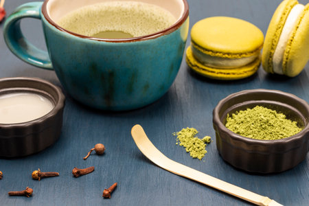 Measuring spoon and green matcha powder. Milk in a bowl. Macaron biscuits and matcha tea in a mug. blue backgroundの写真素材