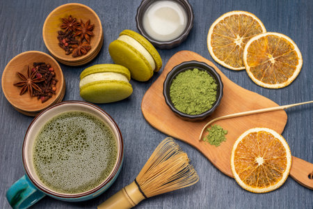 Measuring spoon and green matcha powder on wooden board. Dry orange slices and spices in bowls. Milk in bowl. Macaron biscuits and matcha tea in mug. blue backgroundの写真素材