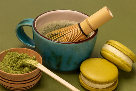 Green matcha powder and measuring spoon in wooden bowl. Green macaroon cookies. Bamboo whisk in mug with matcha tea. green backgroundの写真素材