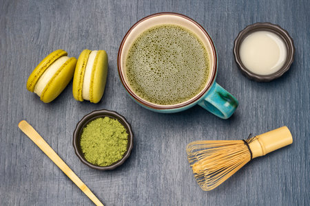Measuring spoon and green matcha powder. Milk in bowl. Macaron biscuits and matcha tea in mug. flat lay. blue backgroundの写真素材