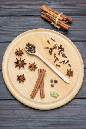 Dry spices on round wooden board. Allspice in spoon, star anise and cloves. Cinnamon sticks are tied into bundle. flat lay. Dark wooden background.の写真素材