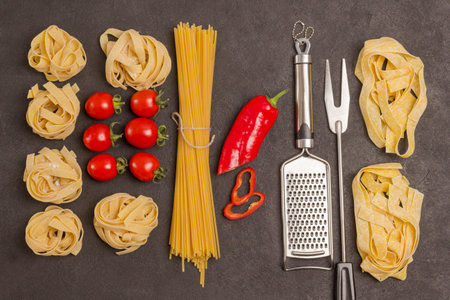 Organic food on black background. Variety of types and shapes of dry Italian pasta with vegetables tomatoes, red peppers, garlic, parsley. Grater and fork. Top viewの写真素材