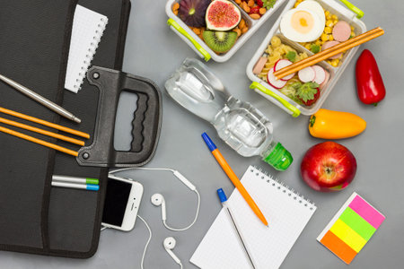 Black briefcase with phone and headphones. Bottle of water, notebooks with pens, lunch box of fruits, berries and nuts Gray background. Flat layの写真素材