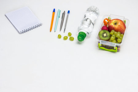 Lunchbox with fruits on table. Notepad Pens, headphones and bottle of water. White background. Copy space. View from aboveの写真素材