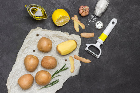 Raw potatoes and one peeled potato on white paper with rosemary, green pepper and lemon. Black background. Flat lay. Copy spaceの写真素材