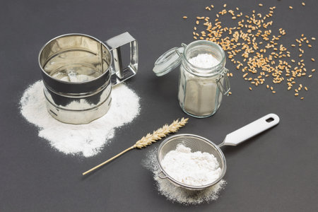 Mug for sifting flour and small sieve with flour, glass jar with flour. Wheat spikelet and wooden...