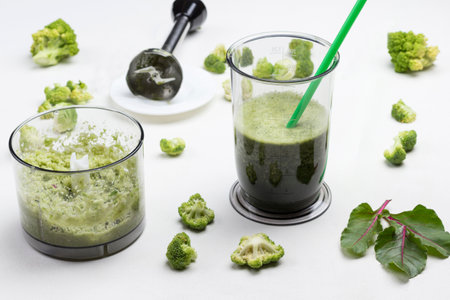 Mugs with green vegetable smoothies. Metal chopper on saucer. Sprigs of rosemary and broccoli on table. White background. Top viewの写真素材