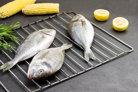 Fresh dorado fish on metal grill. Corn cobs and lemon. Black background. View from aboveの写真素材