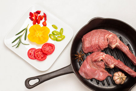 Veal meat on bone, garlic and star anise in pan. Sliced tomatoes and peppers on white plate. Sprig of rosemary and parsley. White background. Flat layの写真素材