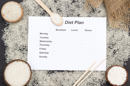 Basmati rice is scattered on table. Round rice in wooden bowl. Bamboo sticks. Rice in wooden spoons. Sheet of paper with diet plan for week. Healthy Diet Plan. Black background. Flat layの写真素材