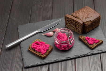 Beet hummus in a glass jar. Beetroot hummus sandwich. Knife and garlic cloves. Loaf of black bread. Dark wooden background. Top viewの写真素材