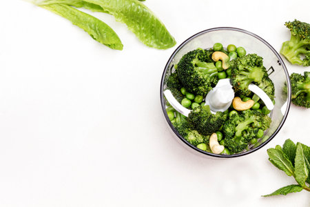 Green peas, broccoli and cashew nuts in blender bowl. Romaine and mint salad on table. Copy space. Top view. White background.の写真素材
