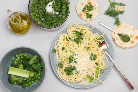 Spaghetti with broccoli paste and fork on ceramic plate. Green peas, mint and broccoli in blender and in bowl. Parmesan cheese on table. Flatbreads with spoon on table. Gray background. Flat layの写真素材
