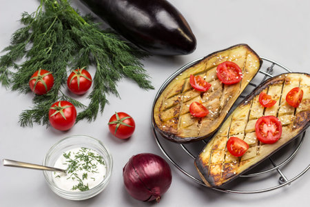 Grilled baked eggplant with tomatoes on metal grill. Dill and tomatoes on table. Sauce in glass bowl. Gray background. Top view.の写真素材