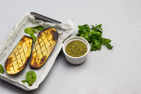 Baked eggplant halves in pallet. Parsley leaves and mung bean in bowl on table. Gray background. Top view. Copy spaceの写真素材